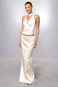 A woman modeling a cream satin two-piece evening gown with a halter-neck crop top and a matching floor-length skirt, set against a neutral gray studio background.