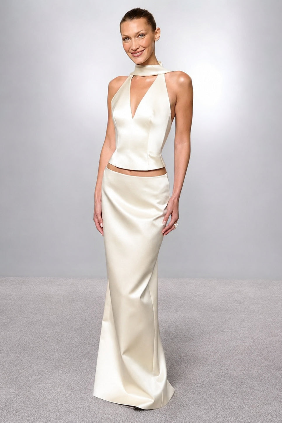 A woman modeling a cream satin two-piece evening gown with a halter-neck crop top and a matching floor-length skirt, set against a neutral gray studio background.