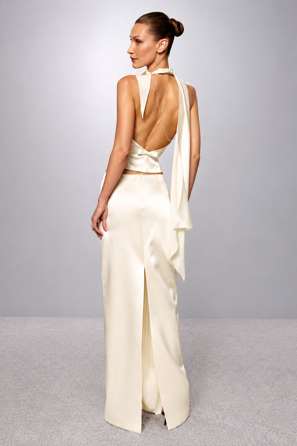 A woman modeling a floor-length ivory satin gown from the back, featuring a high open back, halter-style neck, draped fabric detail, and a front slit, set against a neutral gray studio background.