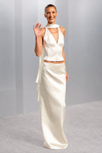 A woman modeling a cream satin two-piece outfit consisting of a sleeveless halter-style top with a deep V-neck and a matching long skirt, standing against a light gray studio backdrop.