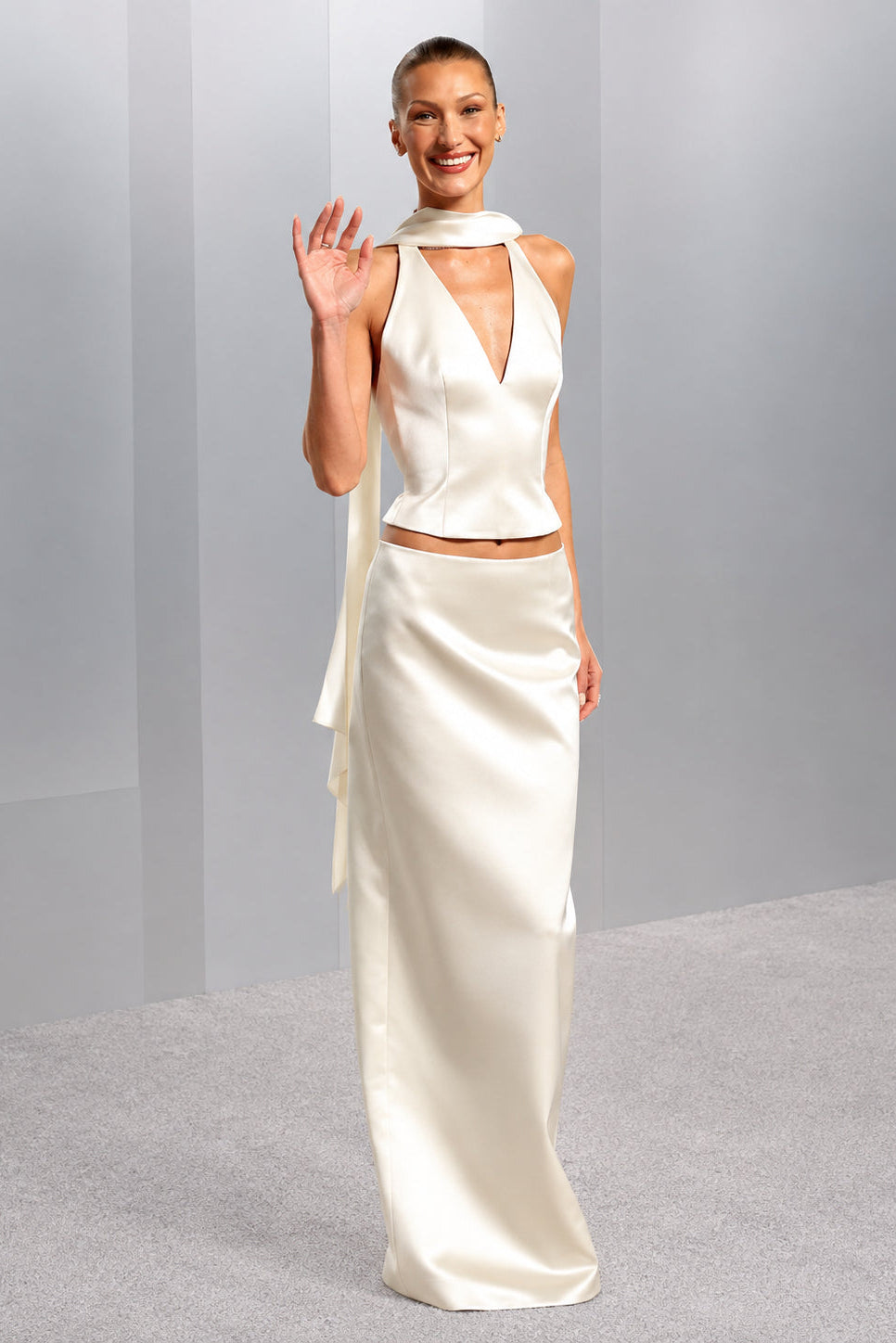 A woman modeling a cream satin two-piece outfit consisting of a sleeveless halter-style top with a deep V-neck and a matching long skirt, standing against a light gray studio backdrop.