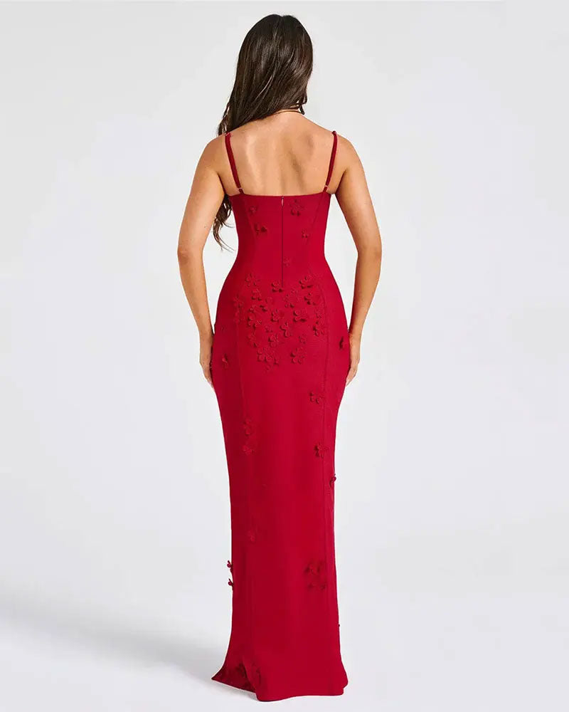 A woman viewed from the back wearing a floor-length red evening gown with thin straps, a fitted silhouette, and floral applique detailing on the back, against a plain light gray studio background.