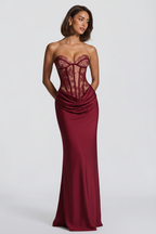 A woman modeling a strapless floor-length gown with a lace corset-style bodice and draped satin skirt in a deep burgundy red, photographed against a light gray studio background.