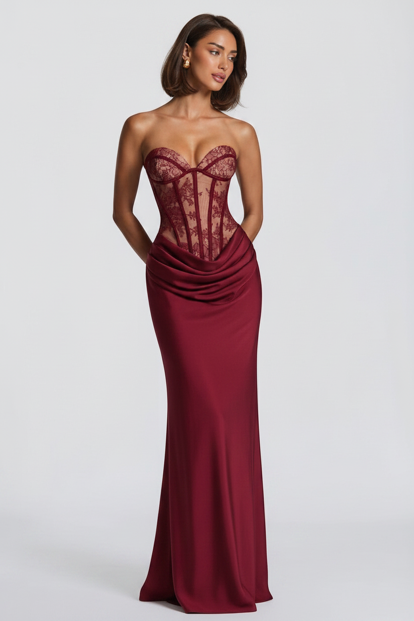 A woman modeling a strapless floor-length gown with a lace corset-style bodice and draped satin skirt in a deep burgundy red, photographed against a light gray studio background.