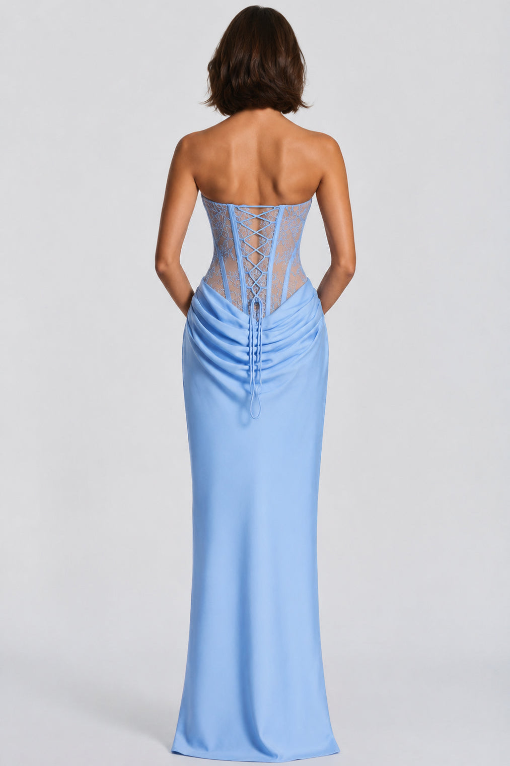 A woman shown from the back wearing a strapless light blue floor-length gown with a lace-up corset-style back and gathered skirt, photographed against a plain light gray studio background.