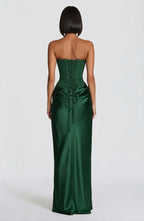 A woman shown from the back wearing a strapless emerald green satin gown with a lace-up corset-style back and gathered skirt, photographed against a plain light gray studio background.