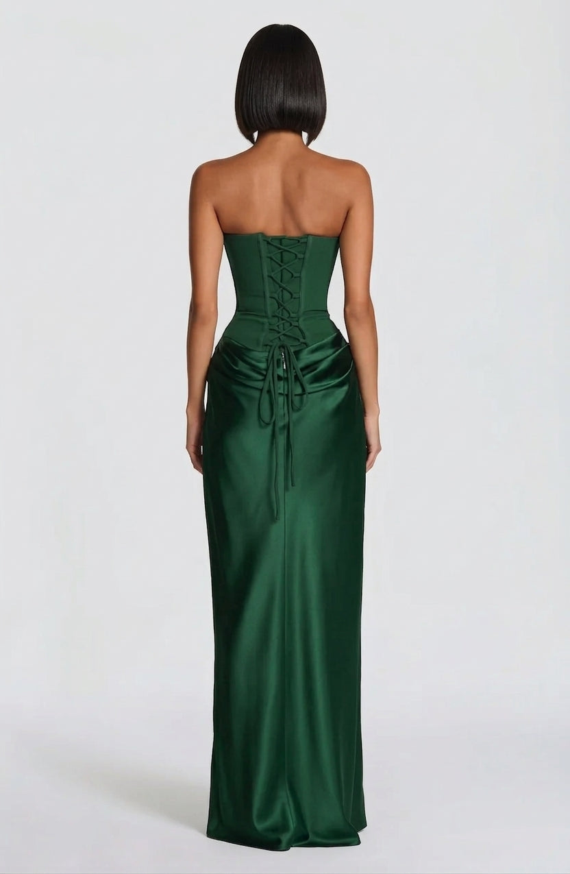 A woman shown from the back wearing a strapless emerald green satin gown with a lace-up corset-style back and gathered skirt, photographed against a plain light gray studio background.