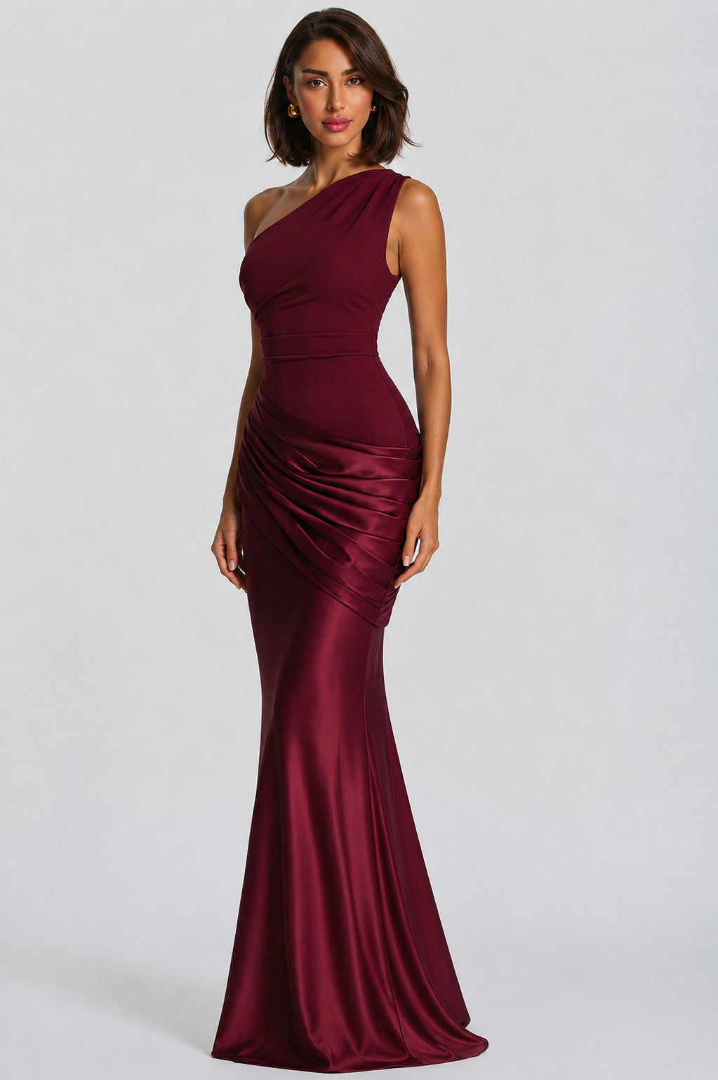 A woman modeling a floor-length, one-shoulder satin gown in deep burgundy against a light gray studio background.