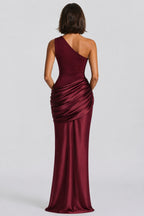 A woman viewed from the back wearing a floor-length one-shoulder satin gown in deep burgundy, with ruched draping at the waist, against a light gray studio background.