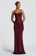 A woman modeling a floor-length halter gown in deep burgundy with gathered draping details at the bodice and skirt, photographed against a light gray studio background.