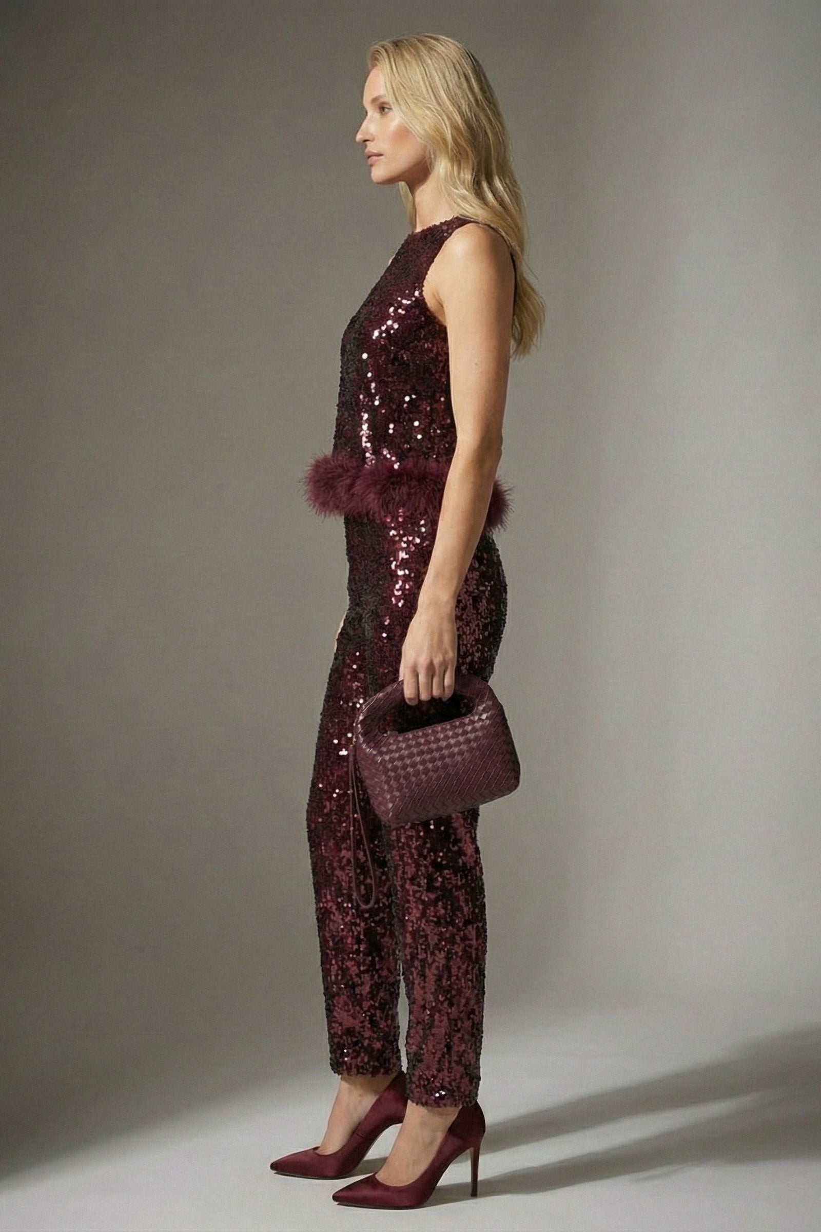 Woman wearing a burgundy sequin jumpsuit with matching shoes and handbag against a gray background