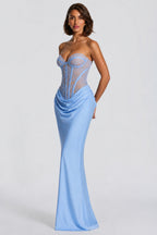 A woman modeling a strapless light blue evening gown with a sheer corset-style bodice and gathered skirt, photographed against a plain light gray studio background.